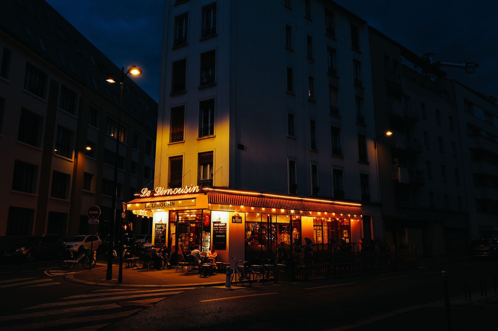 A chauffeur waiting beside a black car outside a restaurant at night